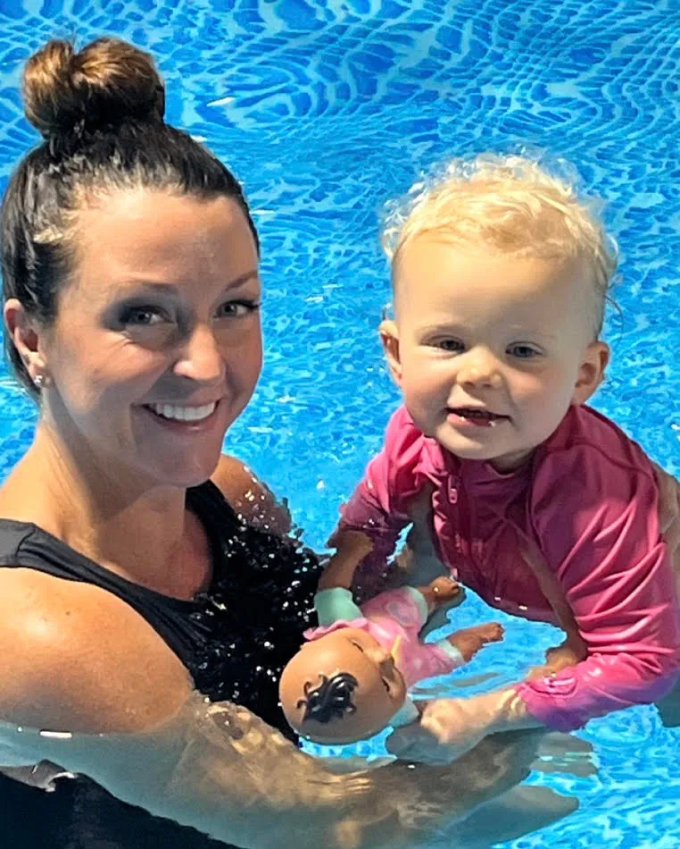 A Tiny Fins instructor holding a smiling toddler in the pool, both looking at the camera